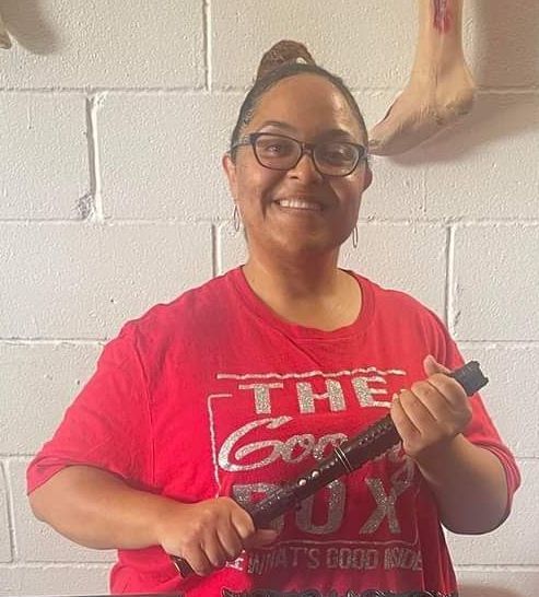 Woman in red shirt smiles, holding a black object. She wears glasses with a bun. White brick wall background.