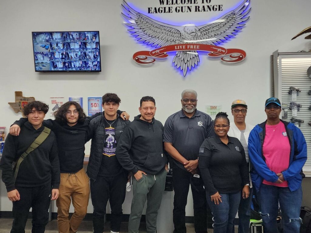 Group of people smiling at Eagle Gun Range, with a sign and surveillance monitors in the background.