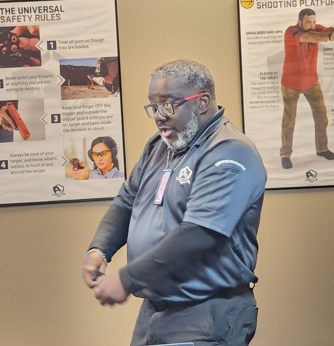 Man in black shirt gesturing during a presentation, with safety posters in the background.