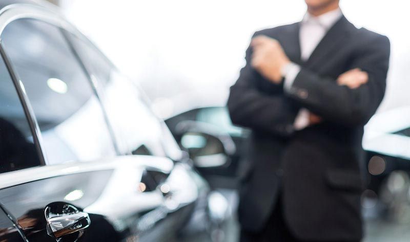Man in suit with arms crossed near a car in a car dealership.