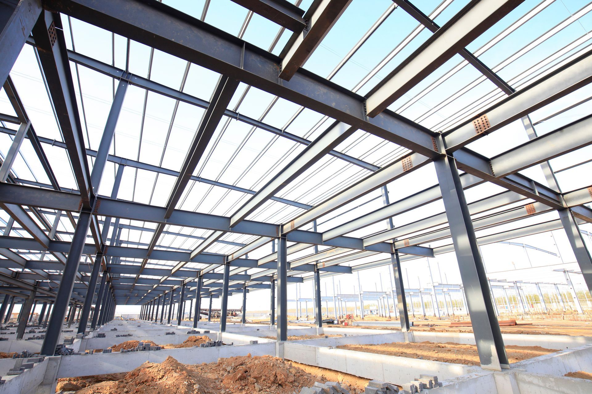 Steel-framed industrial building under construction with active site prep on uneven ground. 