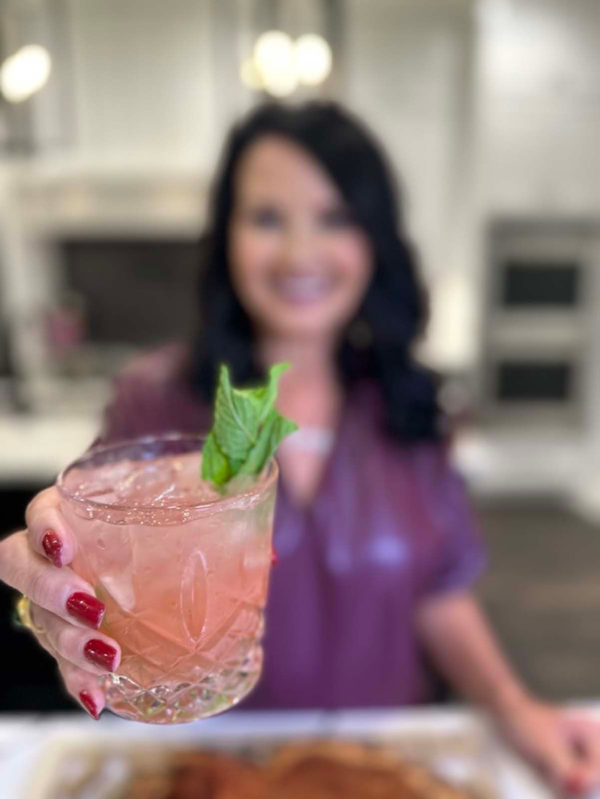 A woman is holding a glass of pink liquid with a mint leaf on top.