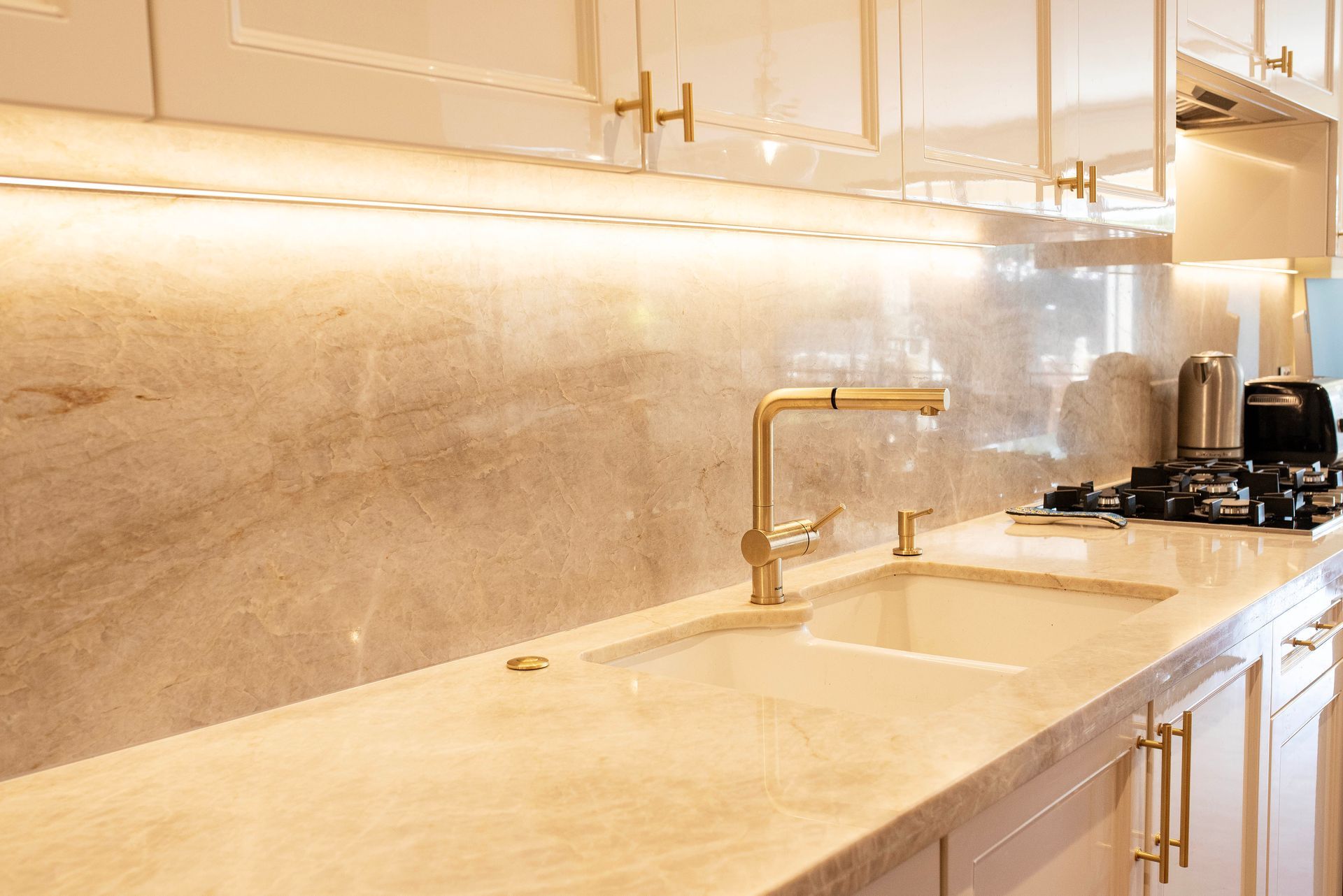 Kitchen sink area with gold faucet, marble backsplash, and under-cabinet lighting.