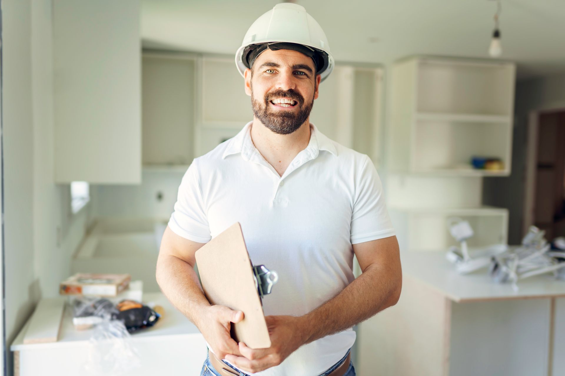 A builder with a white hard hat holding a clipboard. A kitchen renovation is behind him.