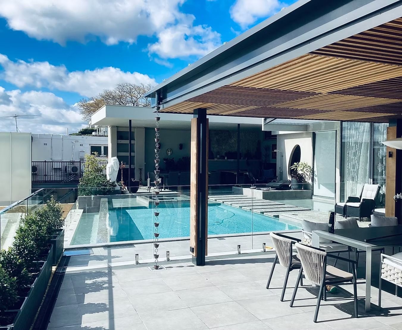 Modern outdoor patio with a wooden slat pergola, overlooking a swimming pool and contemporary house.