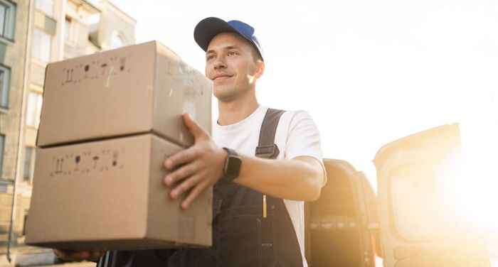 Delivery person carrying two cardboard boxes, sunny outdoor setting.