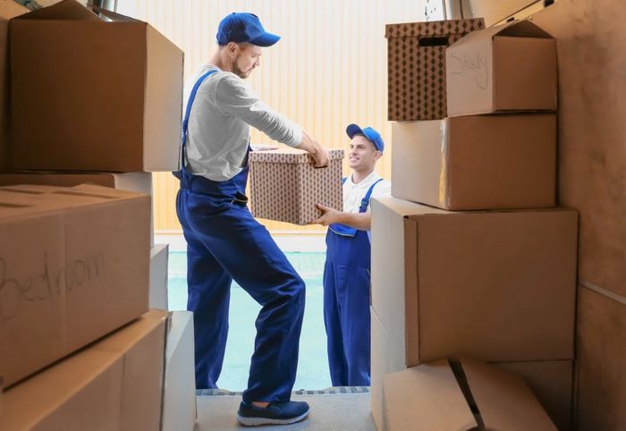 Movers loading boxes into a truck, wearing blue jumpsuits and caps.