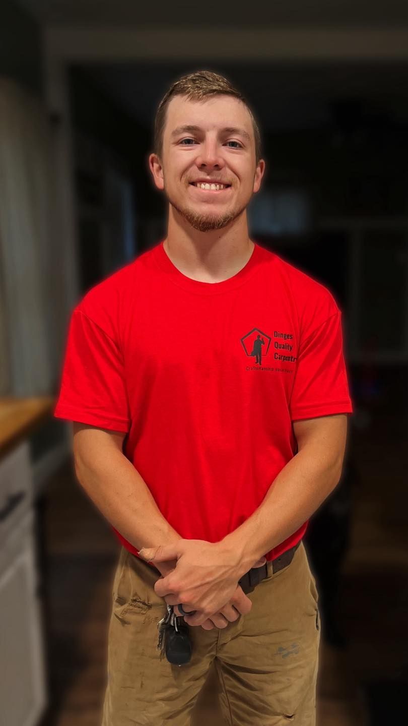 A person wearing a red t-shirt and tan pants, smiling while standing with hands clasped in a dimly lit indoor setting.