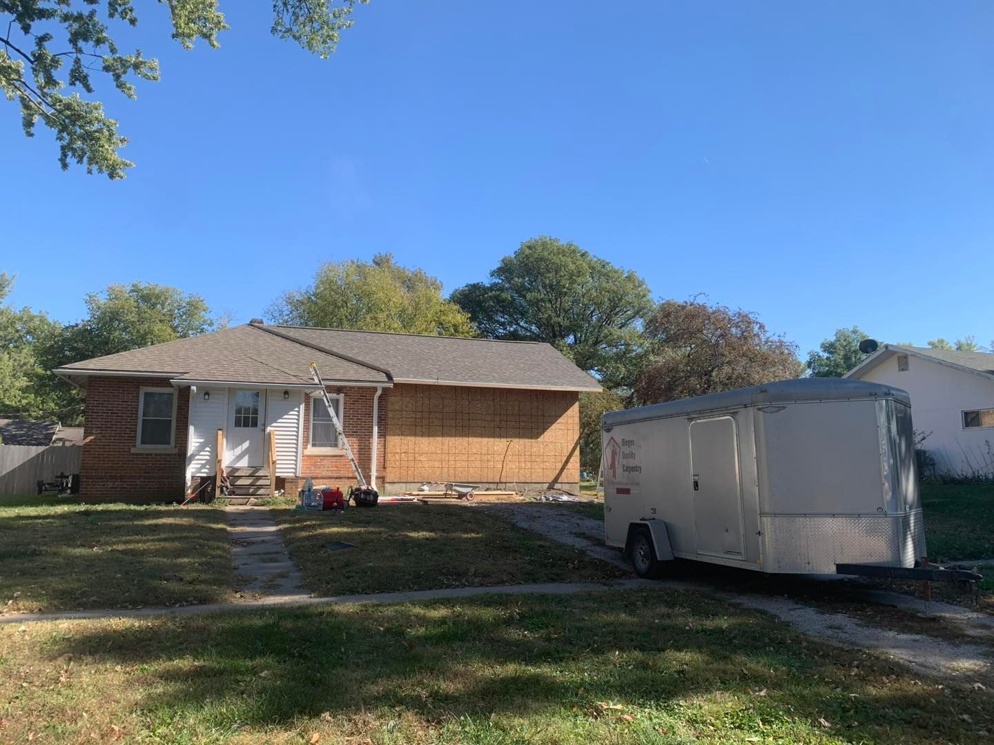 A single-story house with plywood siding under repair sits on a lawn next to a silver cargo trailer on a sunny day.