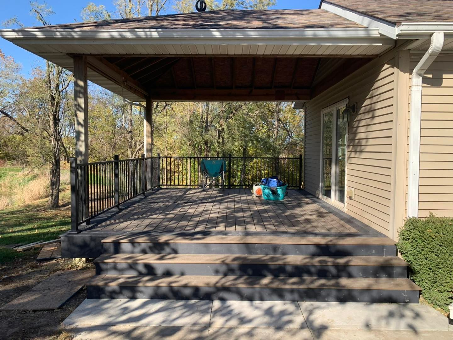 A covered wooden deck with stairs, black railings, and glass sliding doors attached to the side of a beige house.