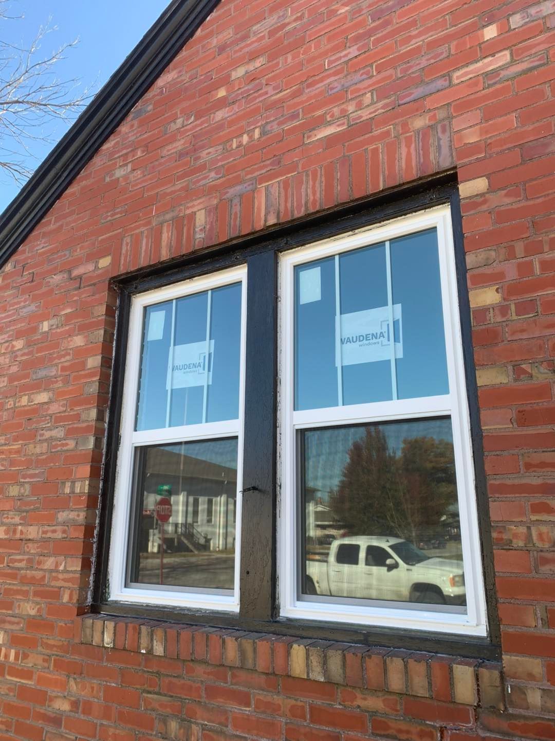 A new, white, double-hung window installed in a red brick exterior wall with a dark-painted wooden frame.
