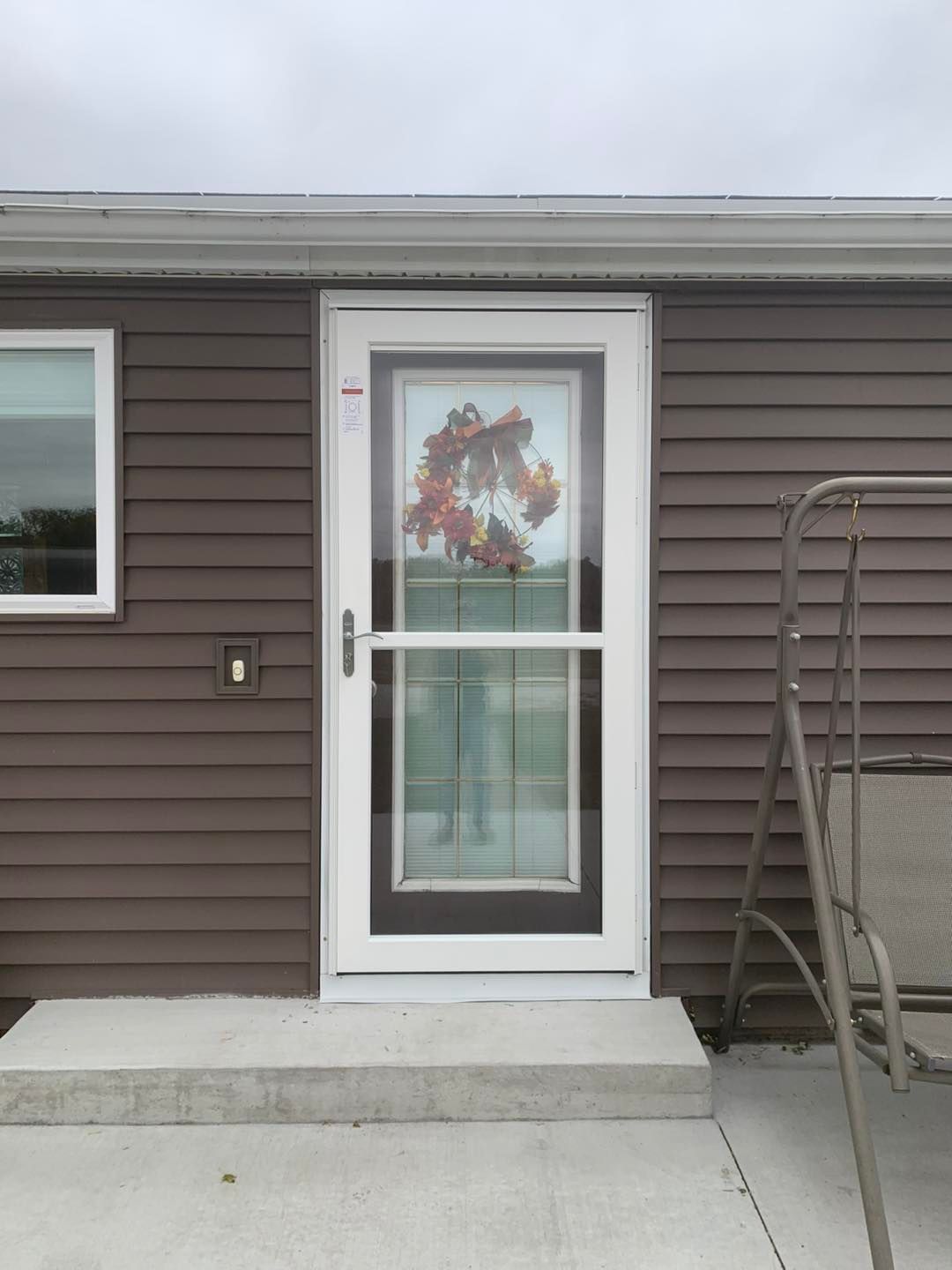 A white storm door with a fall wreath, installed on a house with dark brown horizontal siding and a concrete step.