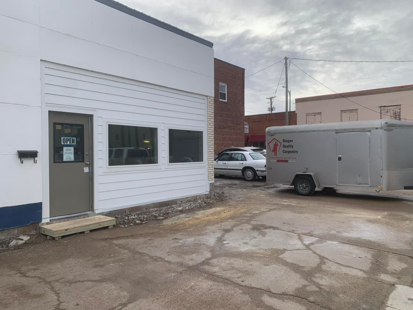 White building exterior with a tan door, small windows, and a gray trailer parked on a cracked paved lot.