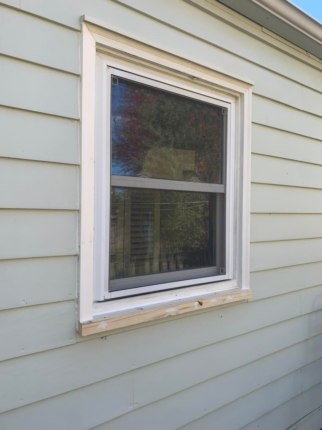 A white-framed window on a light green, horizontal-siding wall with an unfinished wood sill at the base.