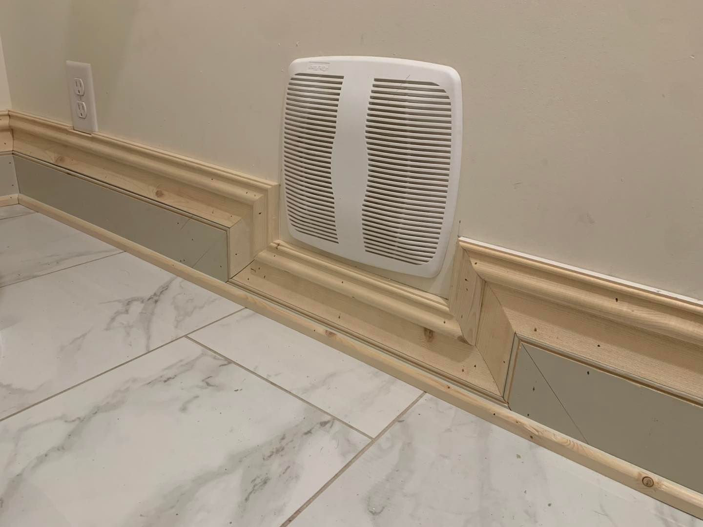 A white wall-mounted vent cover surrounded by decorative tan trim molding installed against marble-patterned floor tiles.