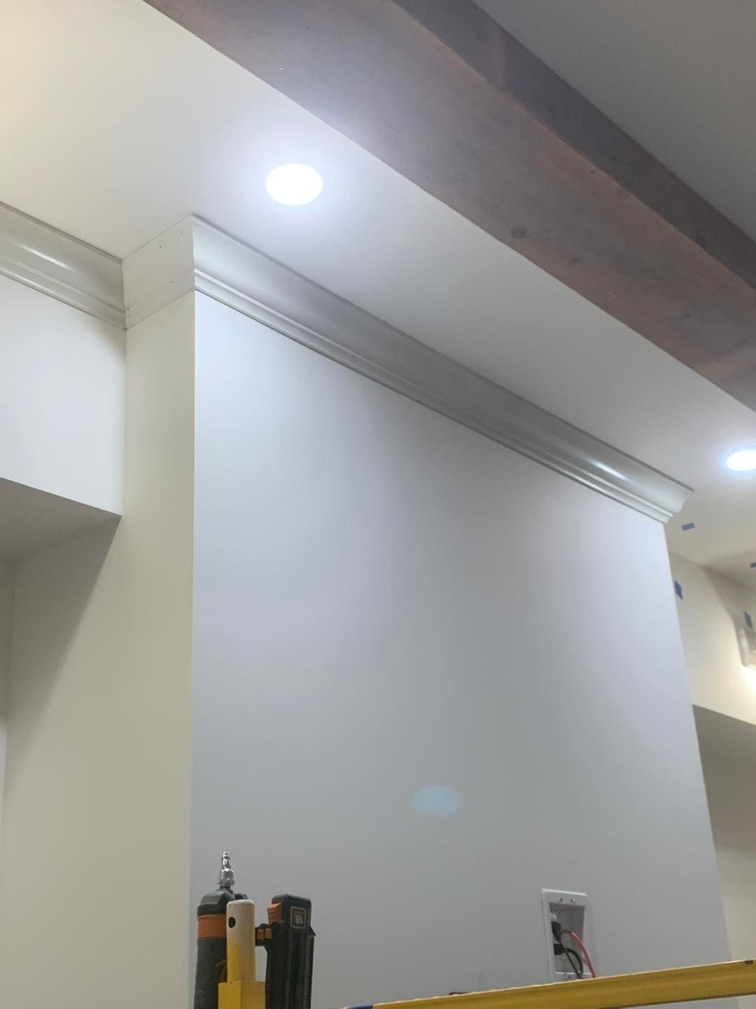 A white wall featuring crown molding at the ceiling, illuminated by recessed lighting with construction tools visible below.