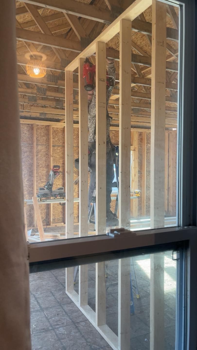 A construction worker framing an interior wall in a room under construction, viewed through a window.