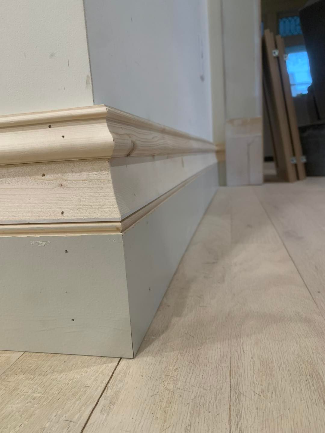 A corner showing light gray baseboard molding topped with a natural wood decorative cap installed on a wooden floor.