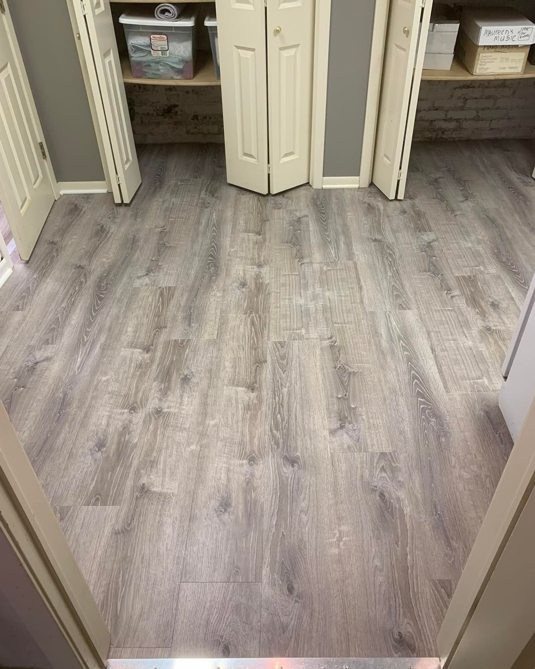 A view into a room with gray wood-look laminate flooring and white closet doors with shelves containing storage bins.