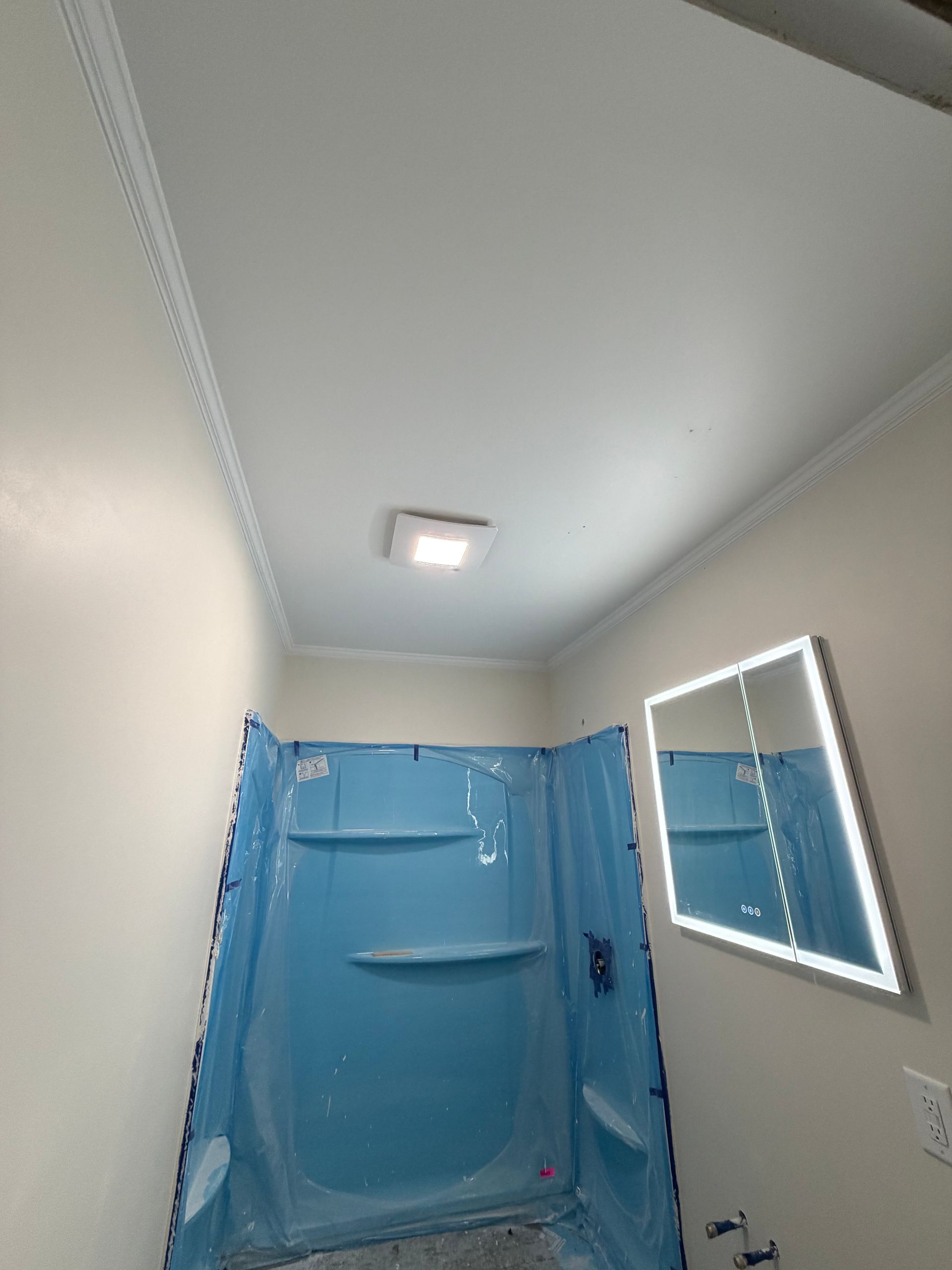 A bathroom showing a blue plastic-covered shower stall, a lit vanity mirror on the wall, and a ceiling light.