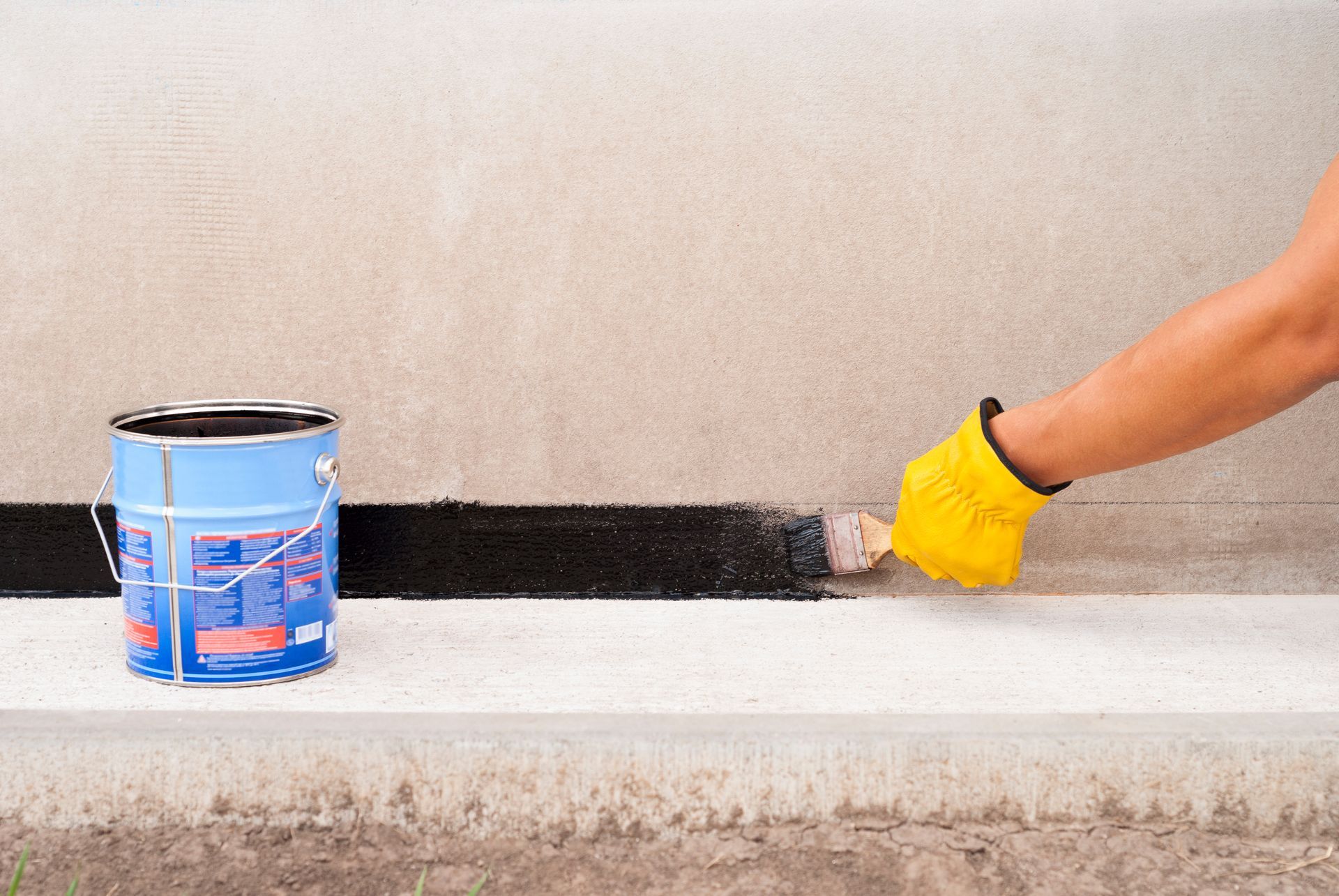 Gloved hand applying black waterproofing to a foundation wall with a paintbrush.