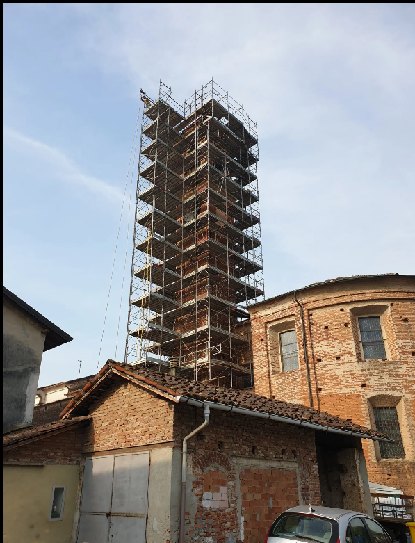 Torre in costruzione, racchiusa tra impalcature, dietro un edificio in mattoni e dei garage.