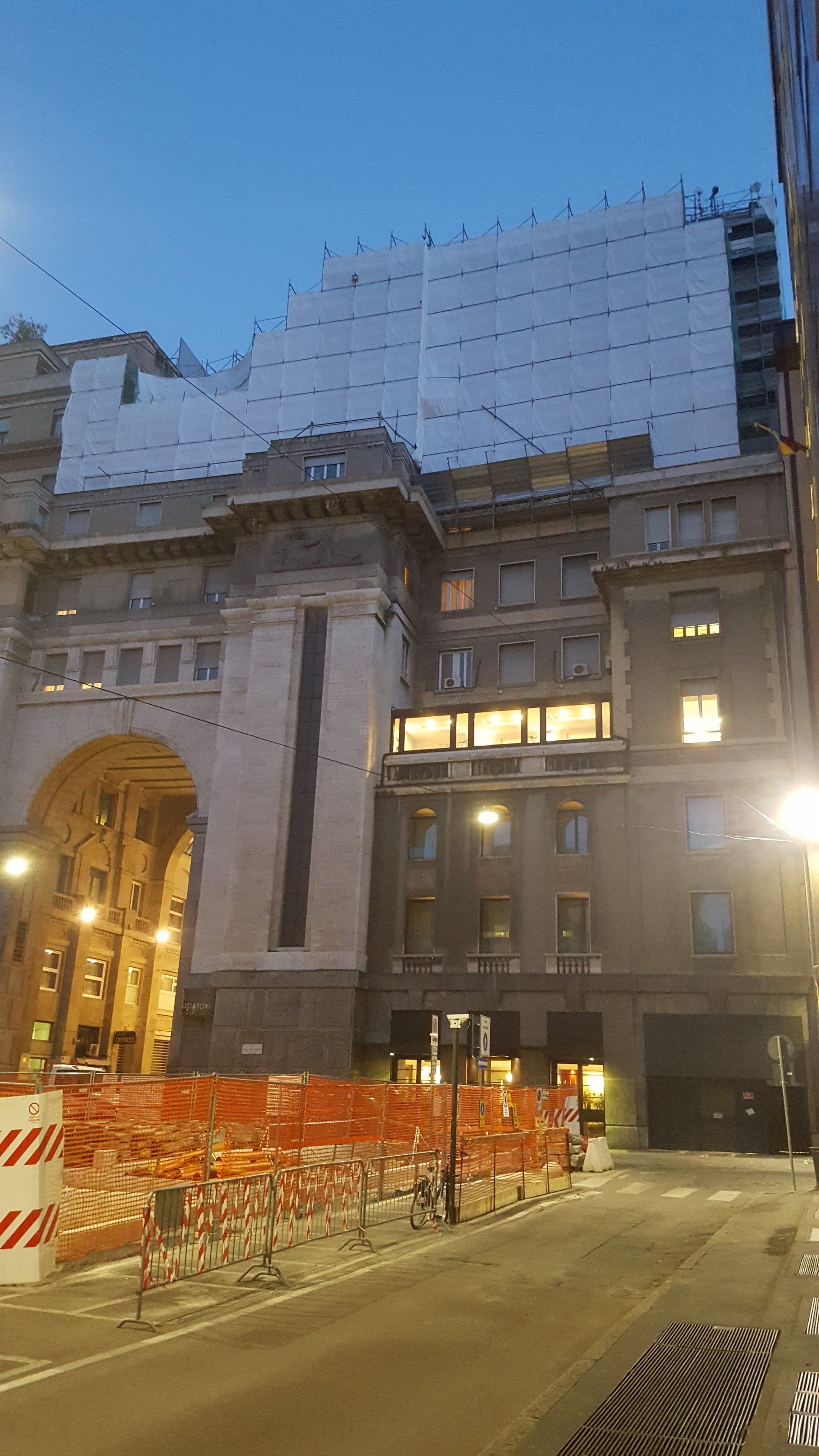 Facciata di un edificio al tramonto, con barriere di sicurezza e impalcature. I colori arancione e bianco sono predominanti.