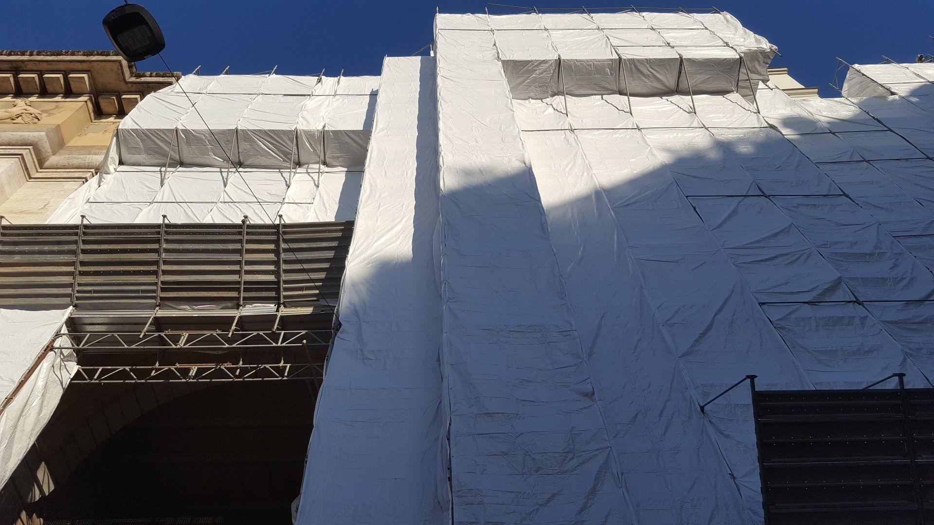 Esterno dell'edificio in fase di ristrutturazione, coperto da teloni bianchi. Impalcature visibili.