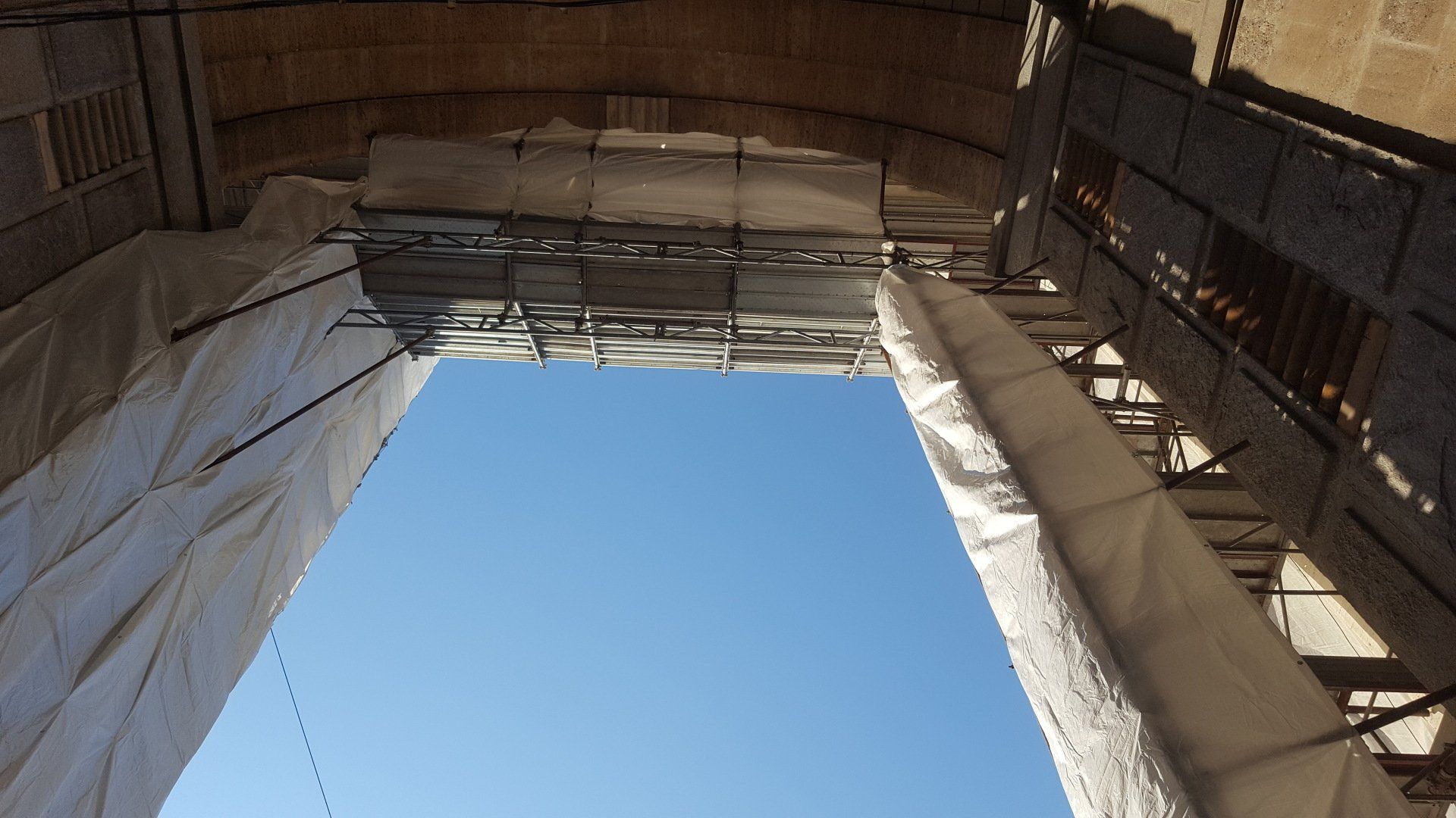 Guardando in alto attraverso un arco, si vedono impalcature avvolte in tessuto bianco e il cielo azzurro.