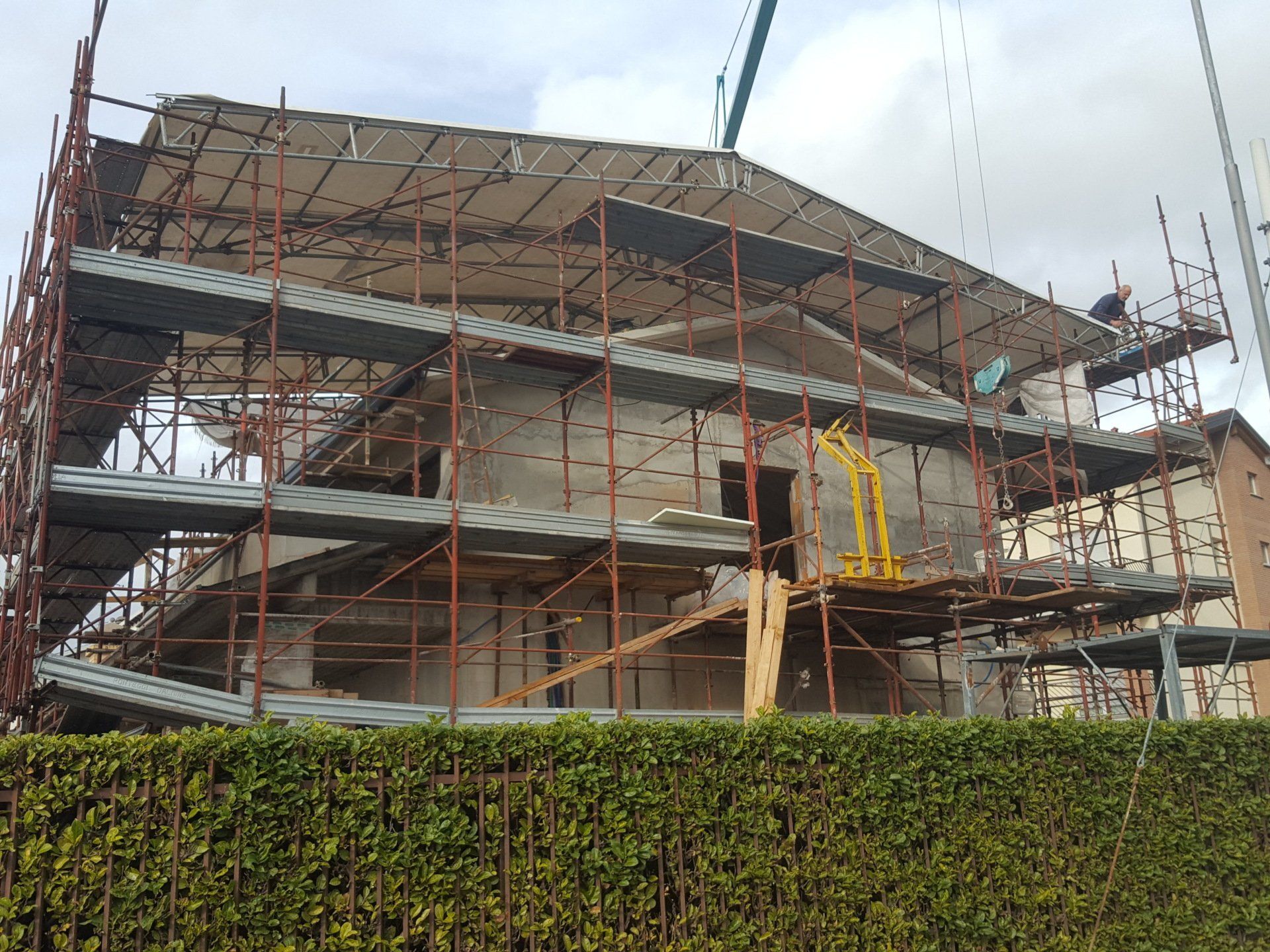 Edificio in costruzione, circondato da impalcature. Struttura grigia, incompiuta, con finestre a vista, siepe verde in primo piano.