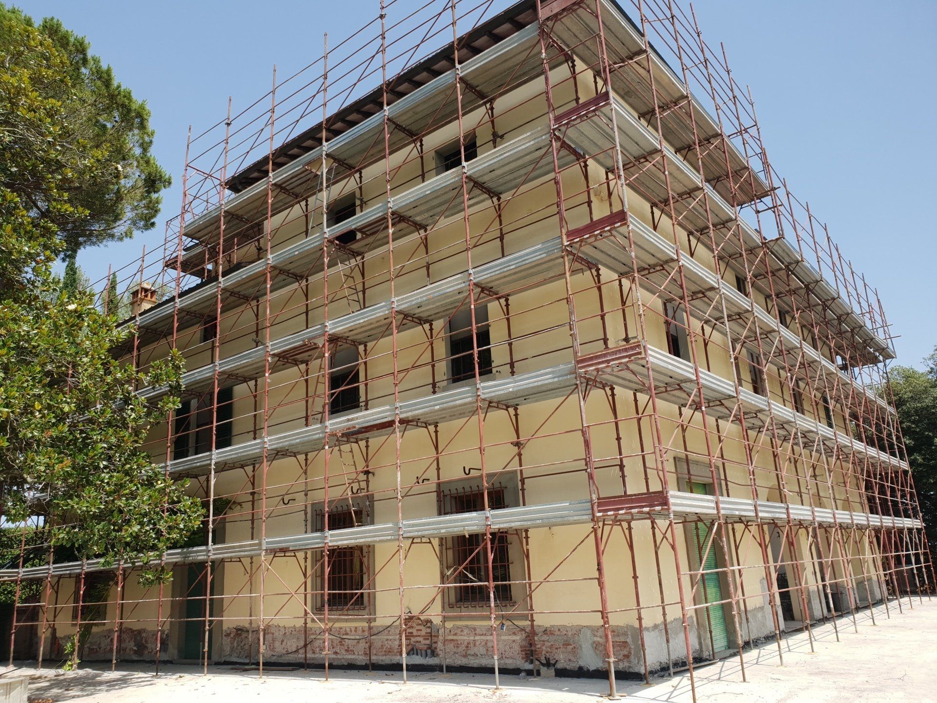 Edificio in ristrutturazione con impalcature; vista esterna. Pareti giallo pallido, finestre marroni, cielo azzurro.