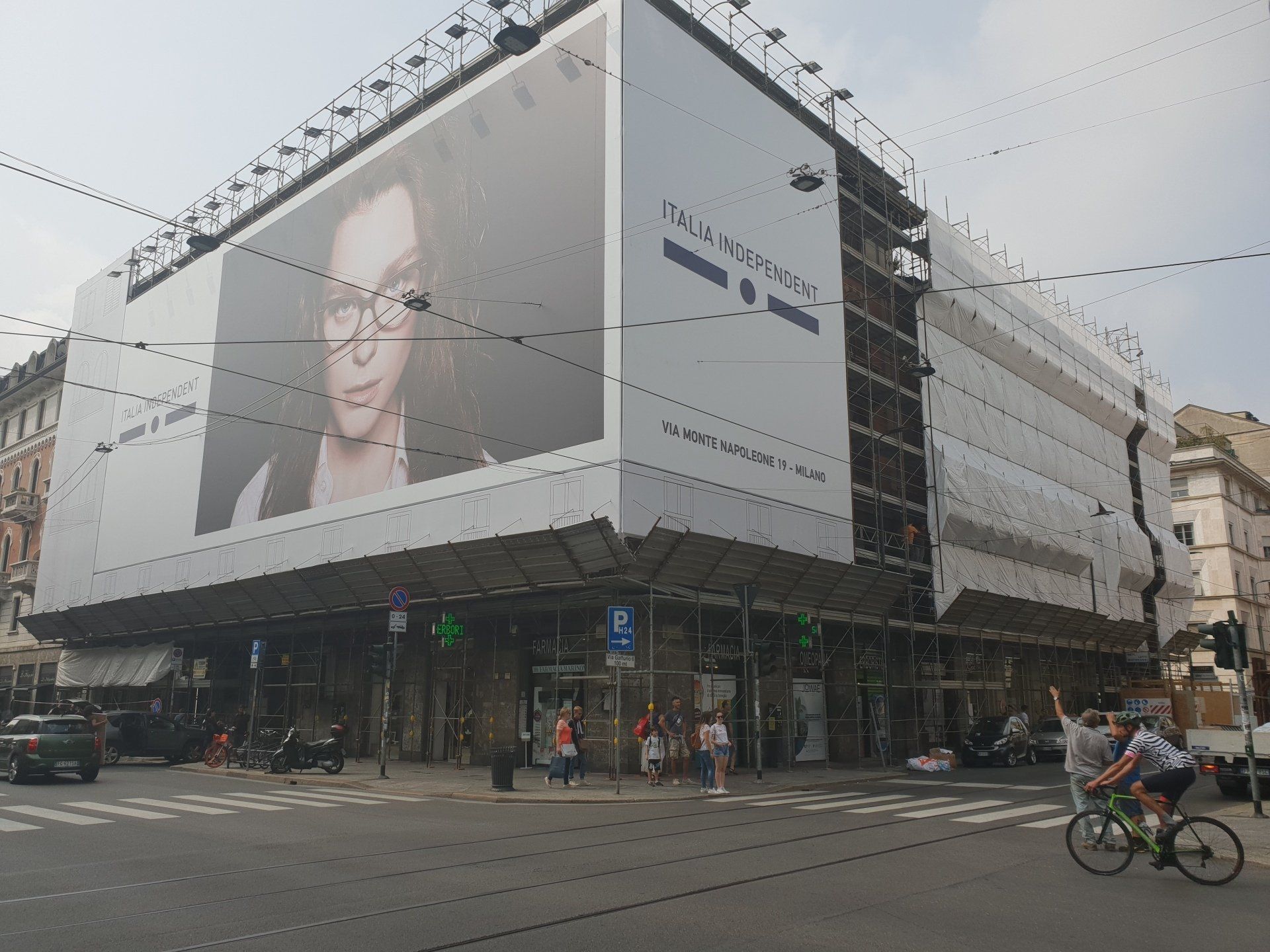 Grande edificio con pubblicità; angolo di strada. Ciclisti, pedoni, veicoli parcheggiati.