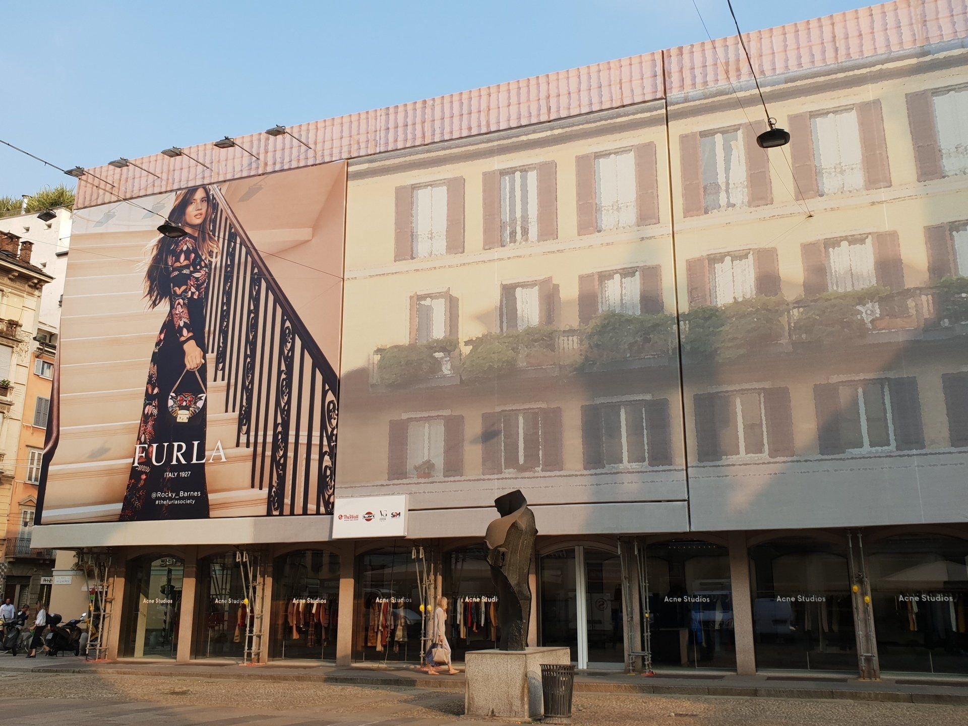 Facciata dell'edificio con grande striscione pubblicitario; vetrine dei negozi sotto.