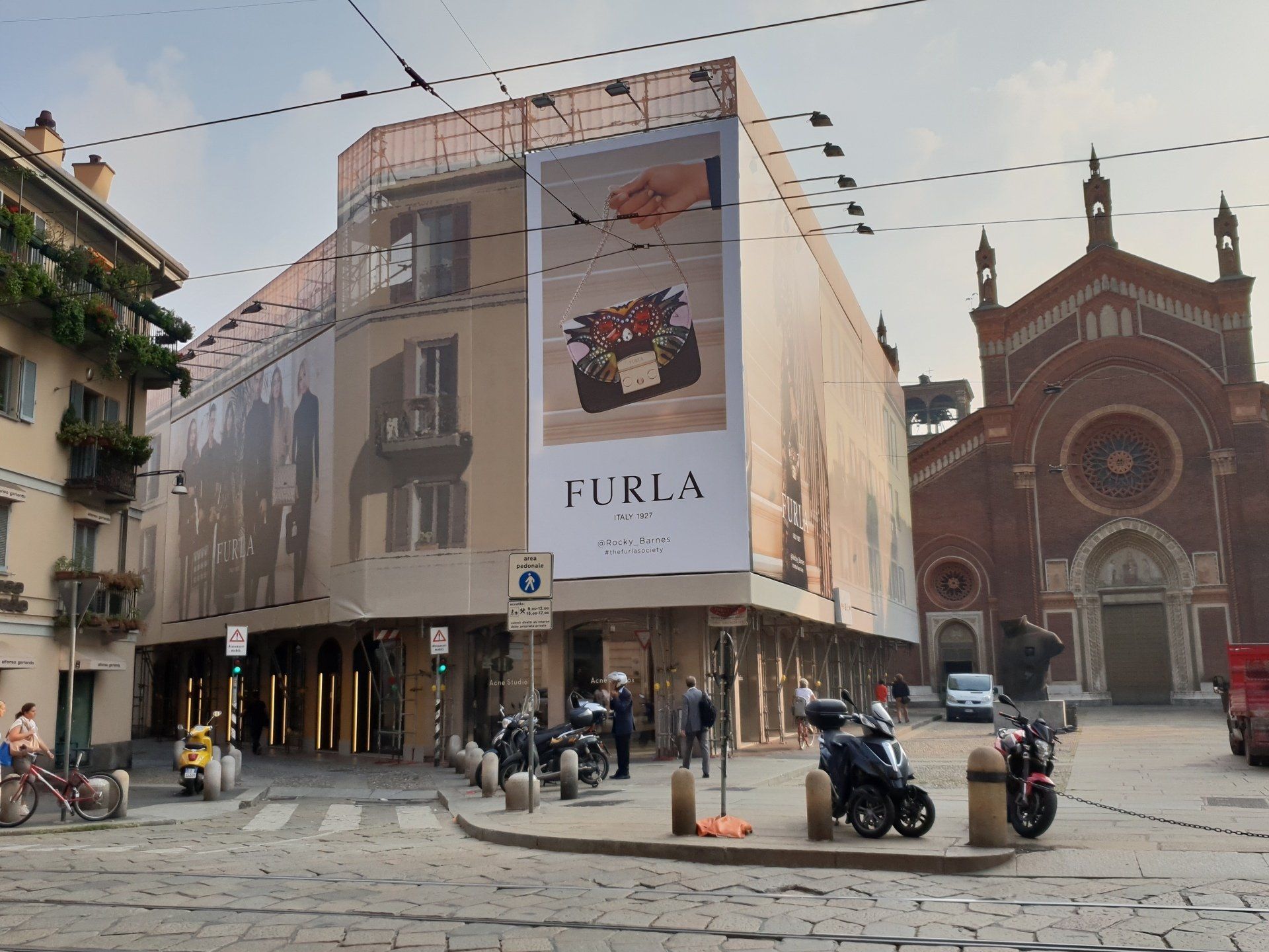 Vetrina di un negozio Furla all'angolo di una città con un grande cartello pubblicitario; sullo sfondo è visibile una chiesa.