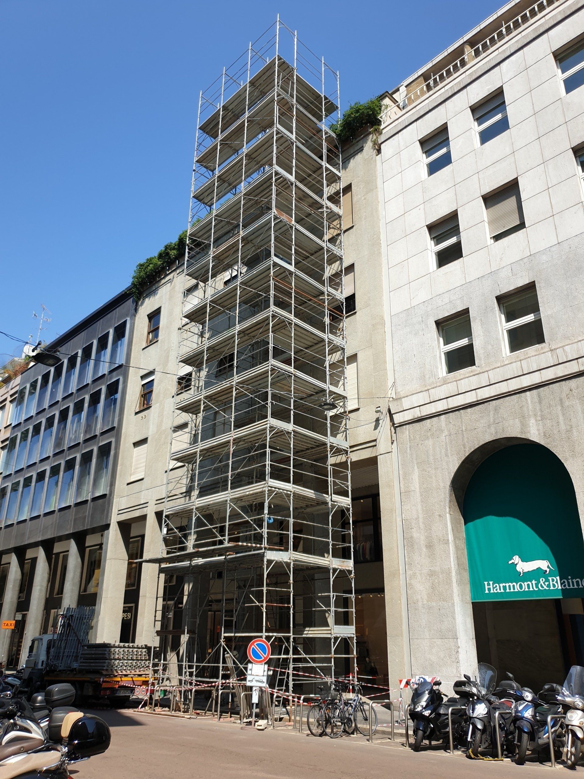 Impalcature accanto a un edificio, probabilmente in riparazione. Biciclette e veicoli parcheggiati in strada. Giornata di sole.