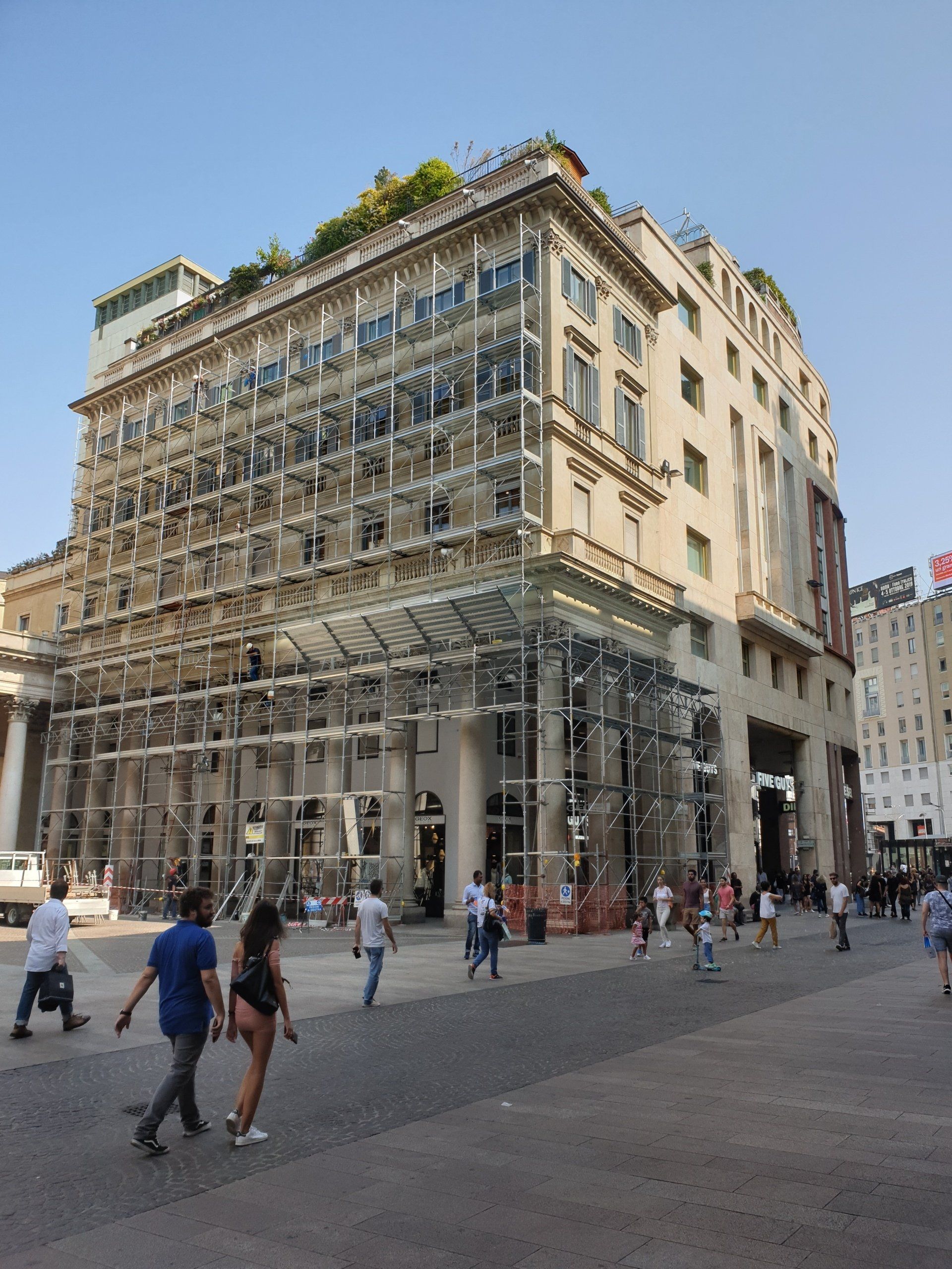 Grande edificio a più piani con facciata in vetro e persone che camminano in una piazza cittadina sotto un cielo limpido.