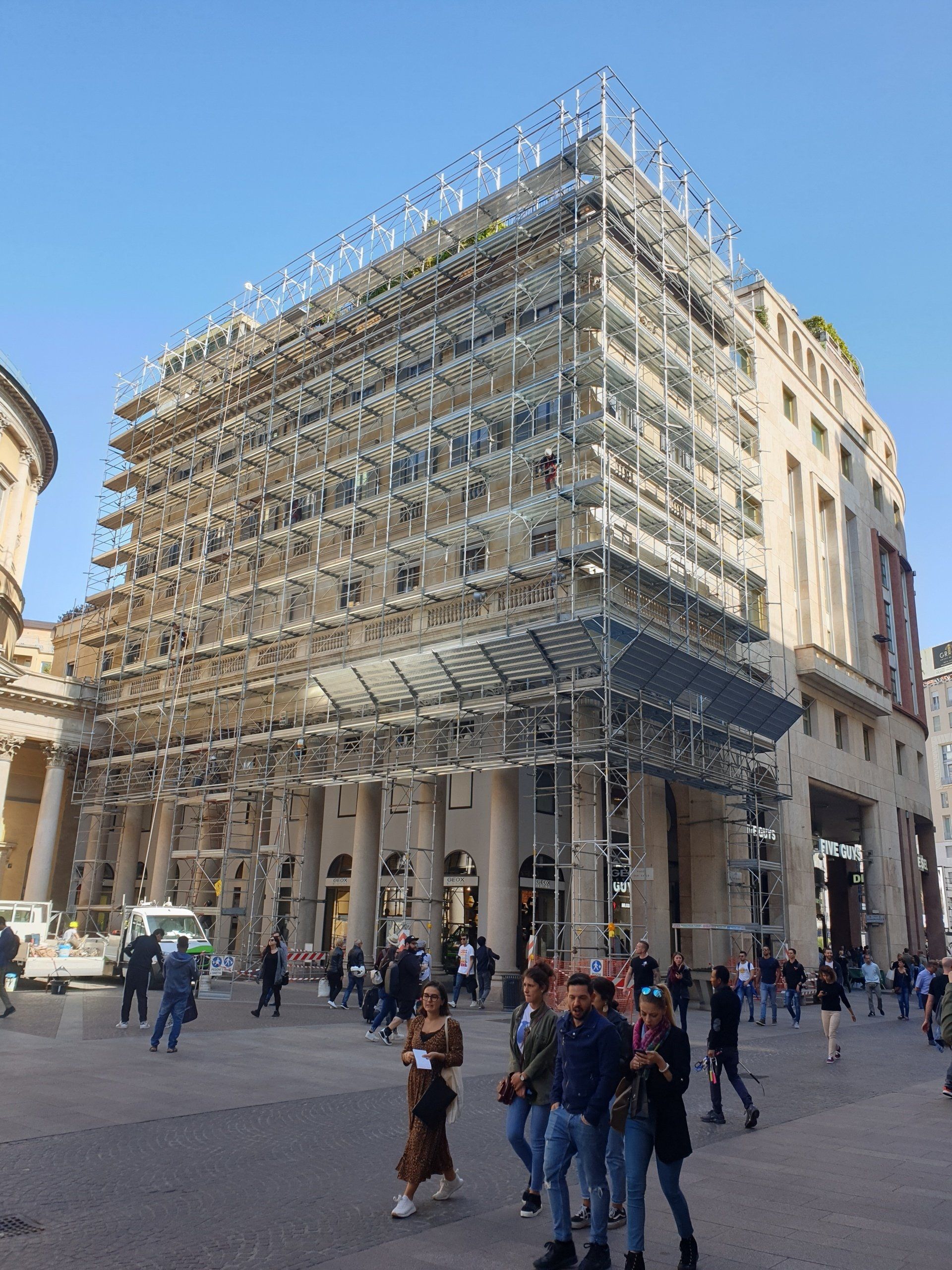 Edificio con impalcature sulla facciata anteriore, persone si radunano in una piazza in una giornata di sole.