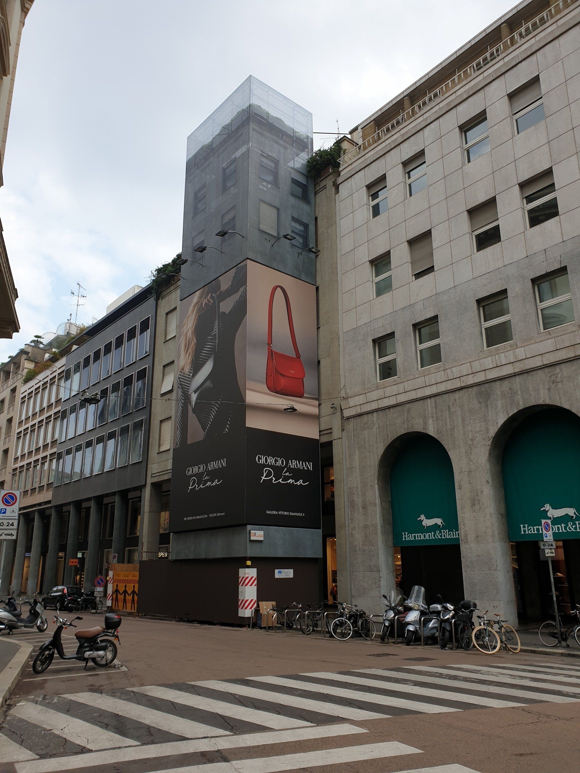 Edifici su una strada cittadina, uno dei quali con un grande cartello pubblicitario raffigurante una borsa rossa. In primo piano si vede un attraversamento pedonale.