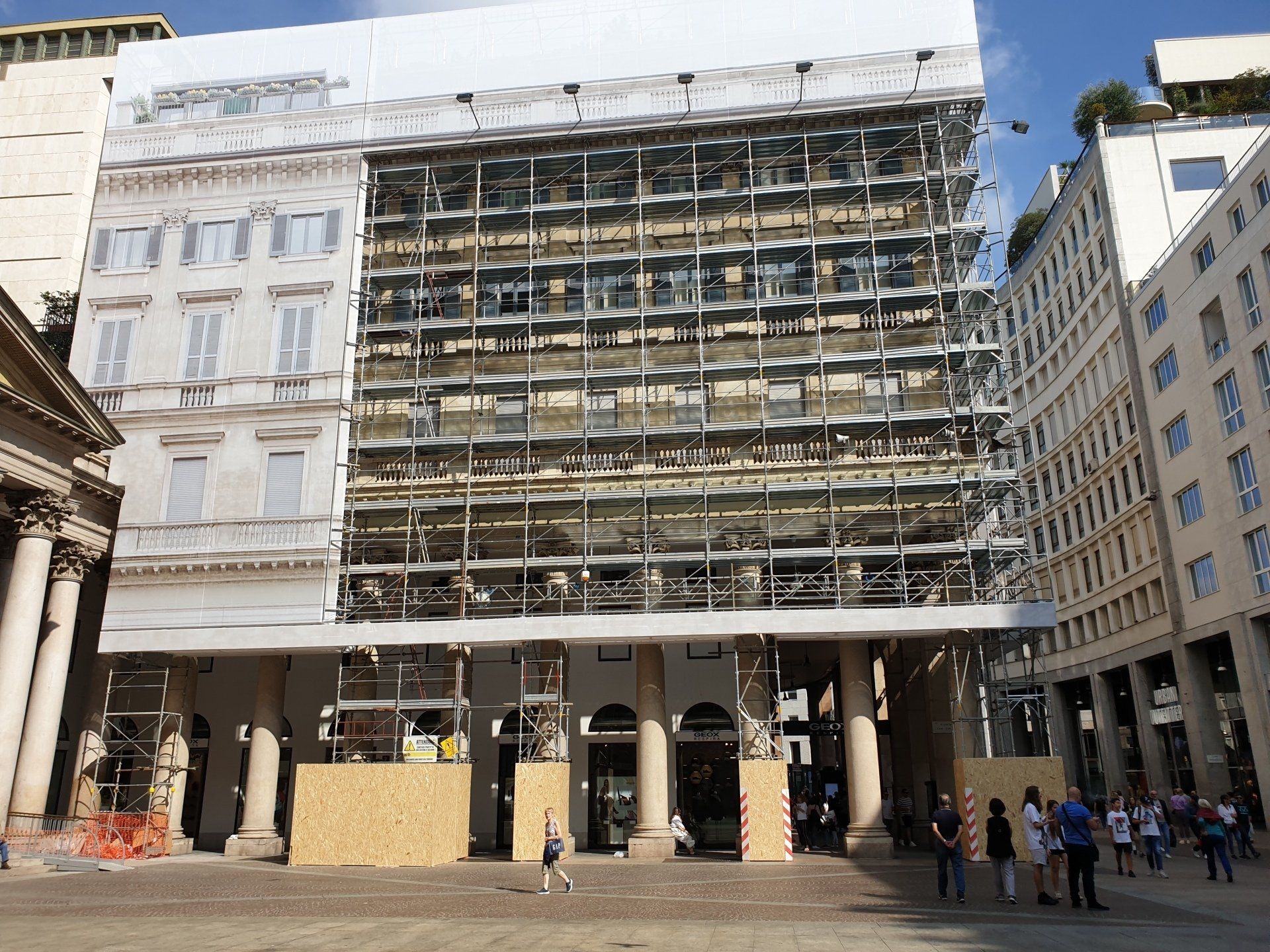 Edificio in fase di ristrutturazione con impalcature e copertura protettiva in una piazza all'aperto con persone.