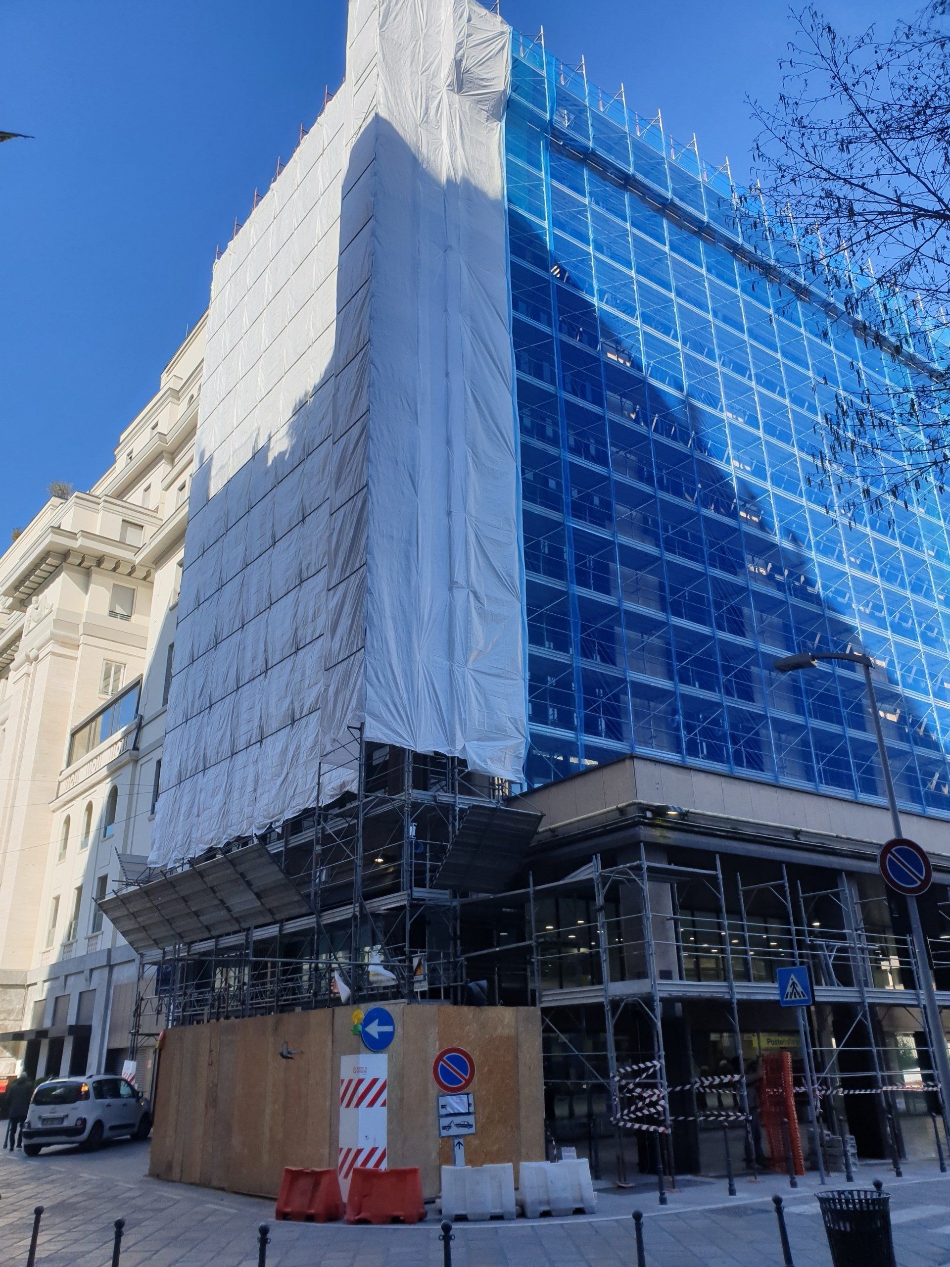 Edificio in costruzione, impalcatura con rete blu, coperture bianche e marroni. Vista dalla strada, luce del giorno.