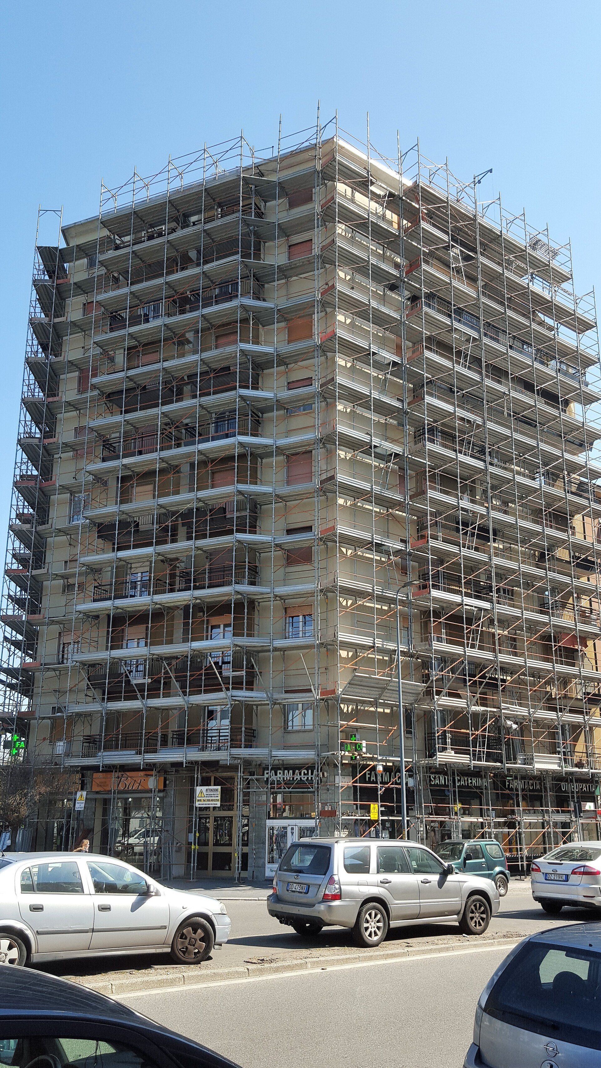 Edificio a più piani con impalcature, veicoli in primo piano, cielo azzurro.