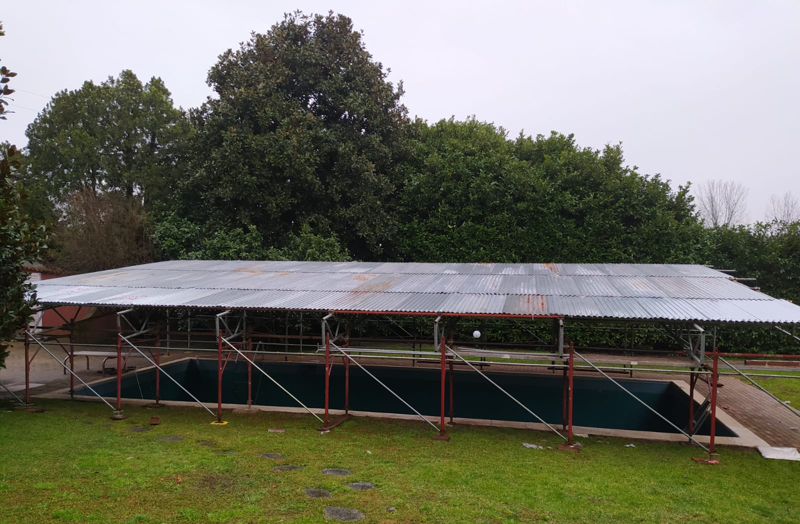 Una piscina coperta da un tetto di lamiera ondulata sostenuto da impalcature rosse; immersa nell'erba verde con alberi sullo sfondo.