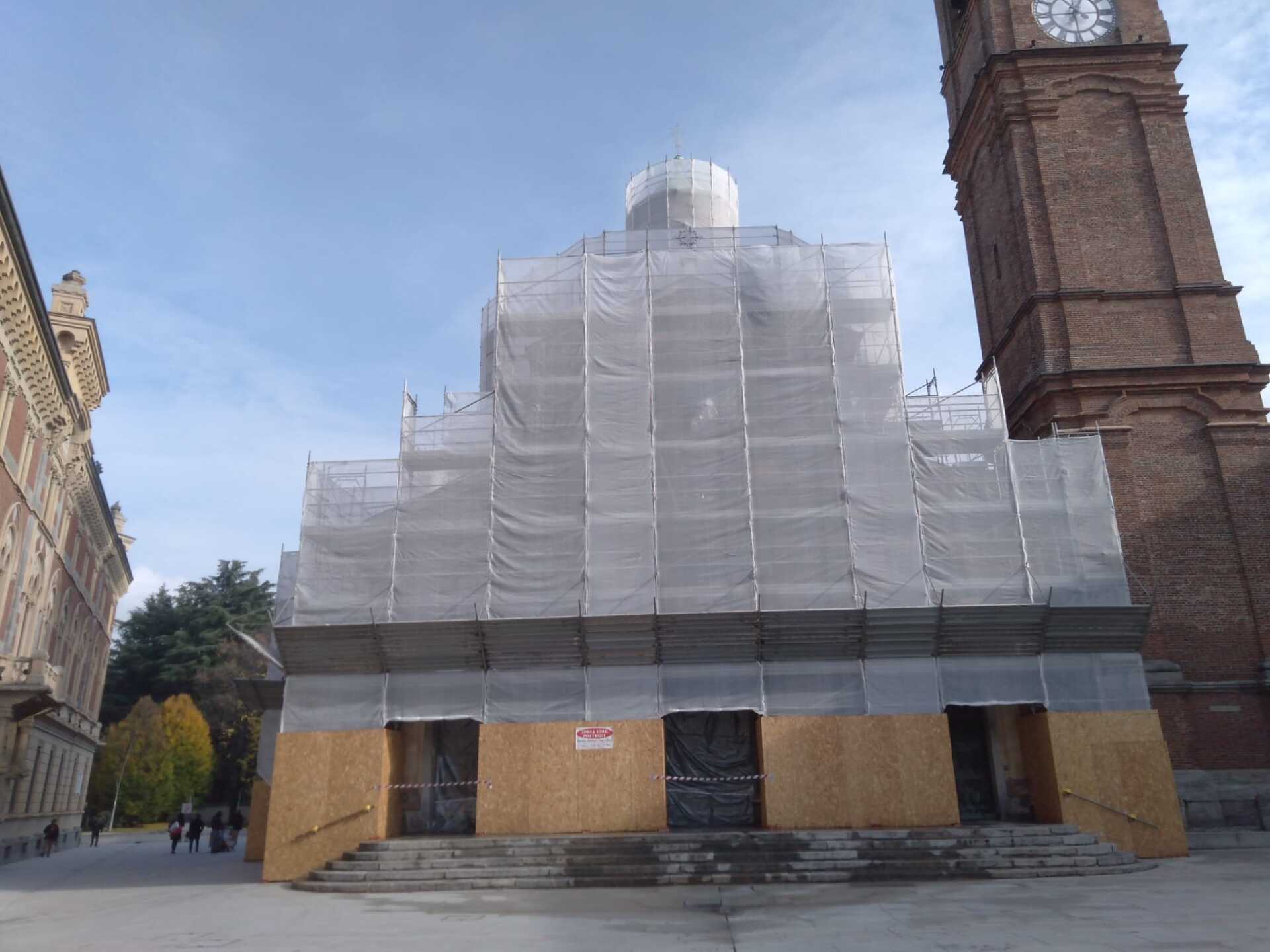 Facciata della chiesa in fase di ristrutturazione, coperta da impalcature e teli protettivi, con una torre dell'orologio sulla destra.