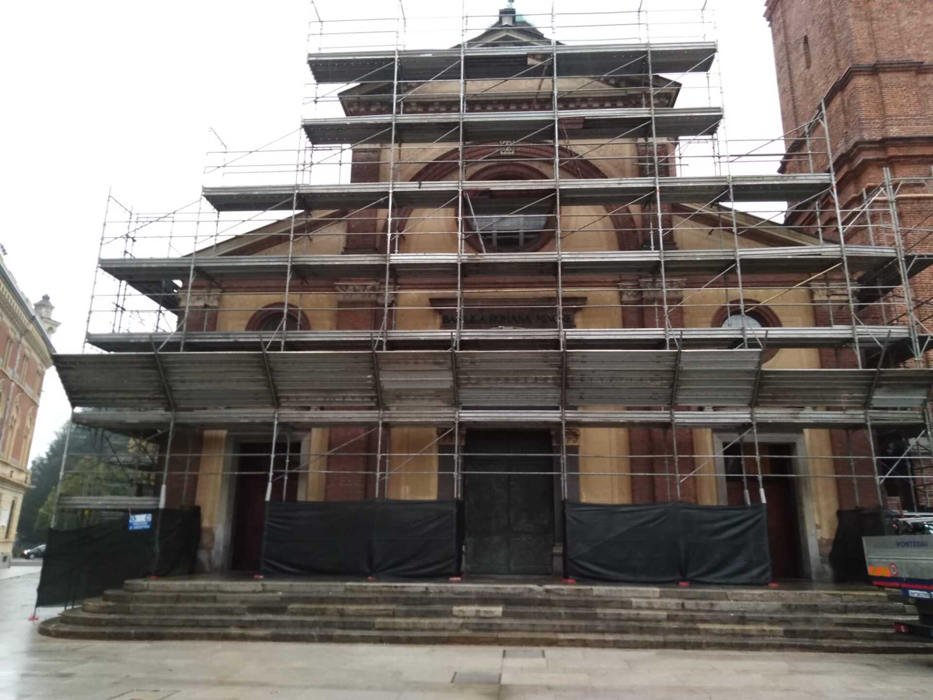 Facciata della chiesa con impalcature, in fase di restauro. Edificio beige e marrone con gradini.