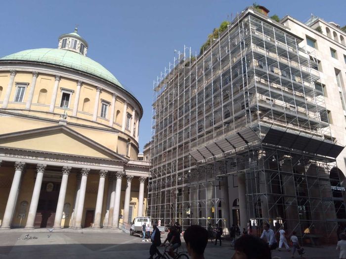 Cupola gialla di una chiesa e edificio sotto impalcature in una piazza urbana piena di gente.