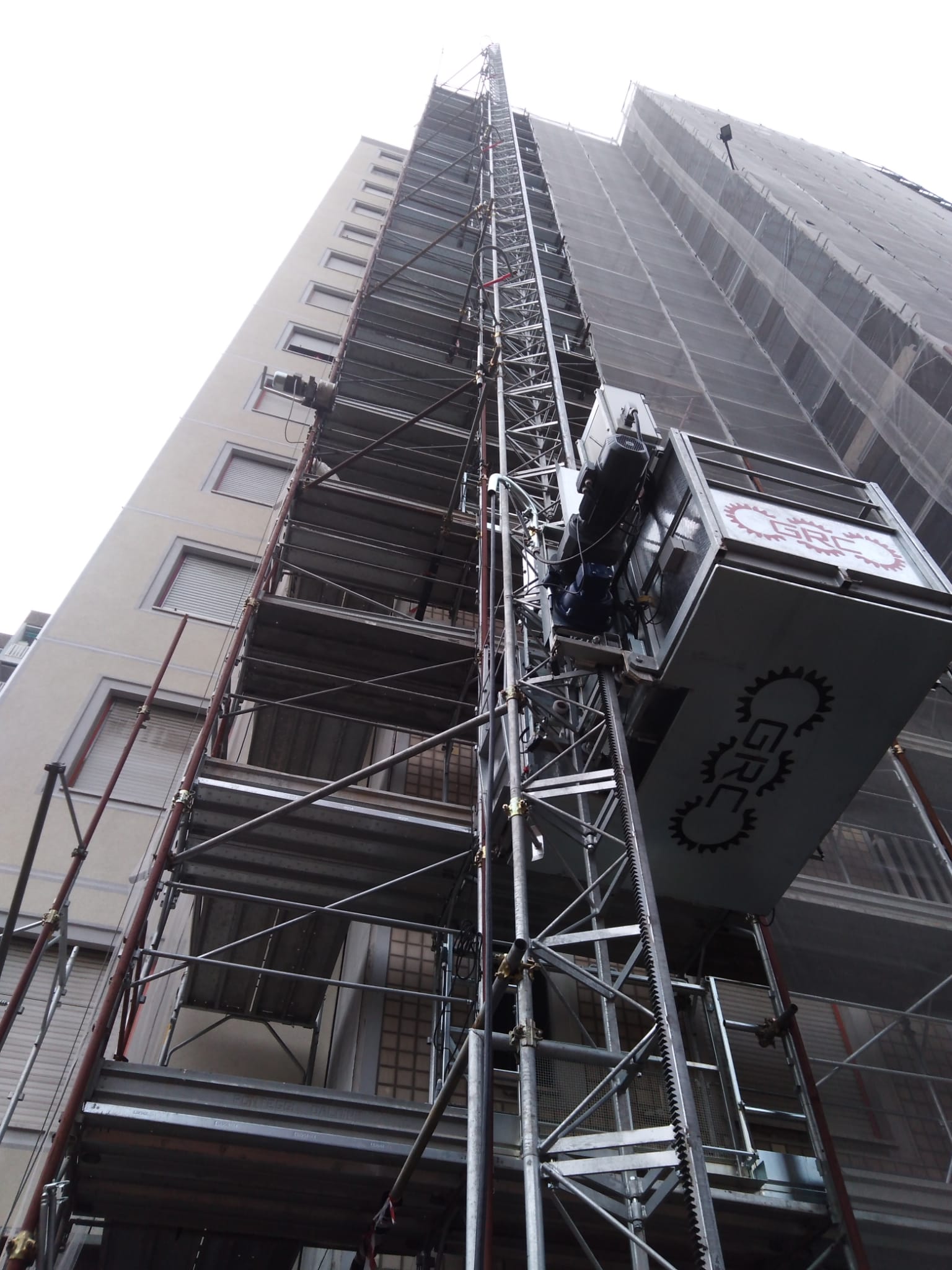La piattaforma dell'ascensore sale sulle impalcature di un alto edificio. Il cielo è coperto.