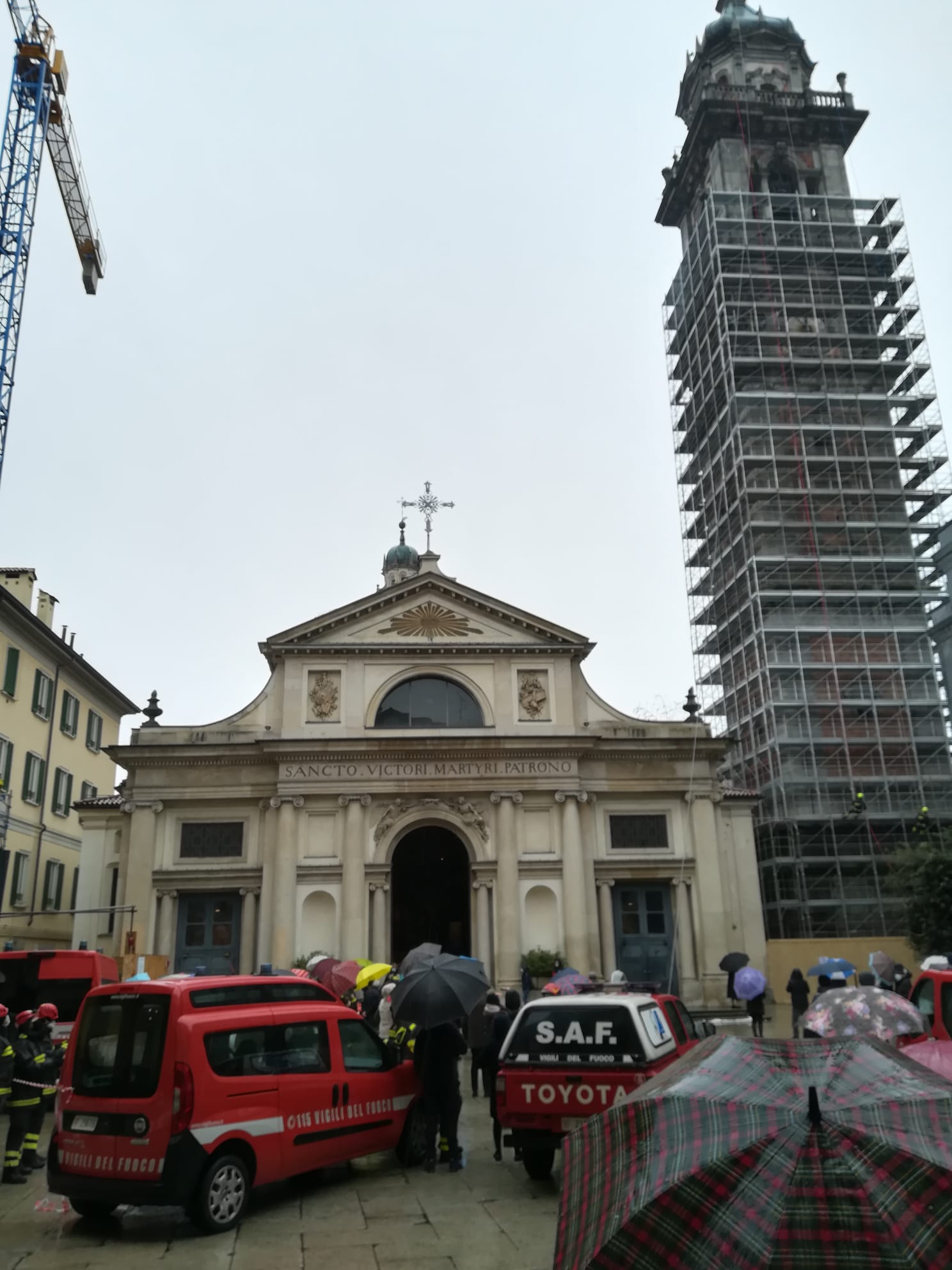 Chiesa con impalcature sulla torre, mezzi di soccorso e persone con ombrelli davanti.