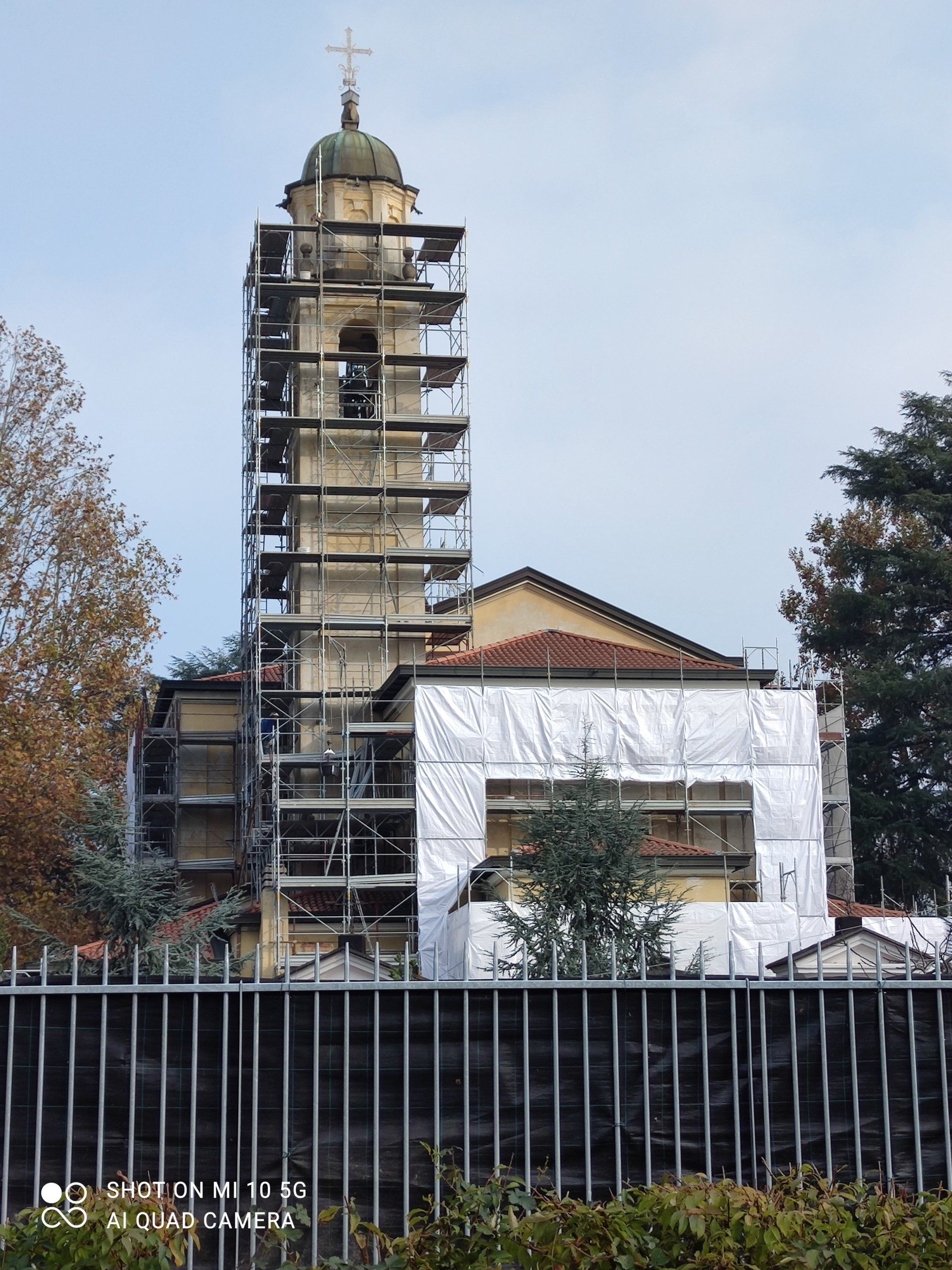 Campanile della chiesa in fase di ristrutturazione, coperto da impalcature, dietro una recinzione metallica.