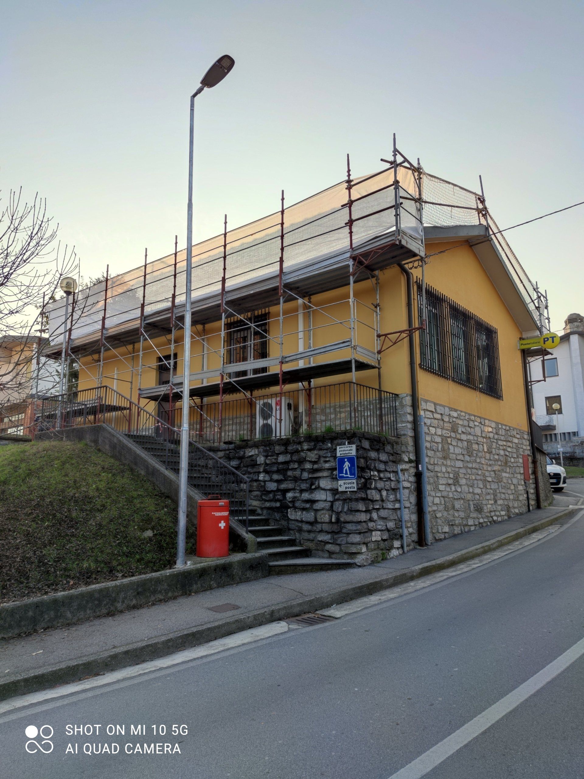 Edificio giallo con impalcature, basamento in pietra e strada. Sono presenti un lampione e una cassetta postale rossa.