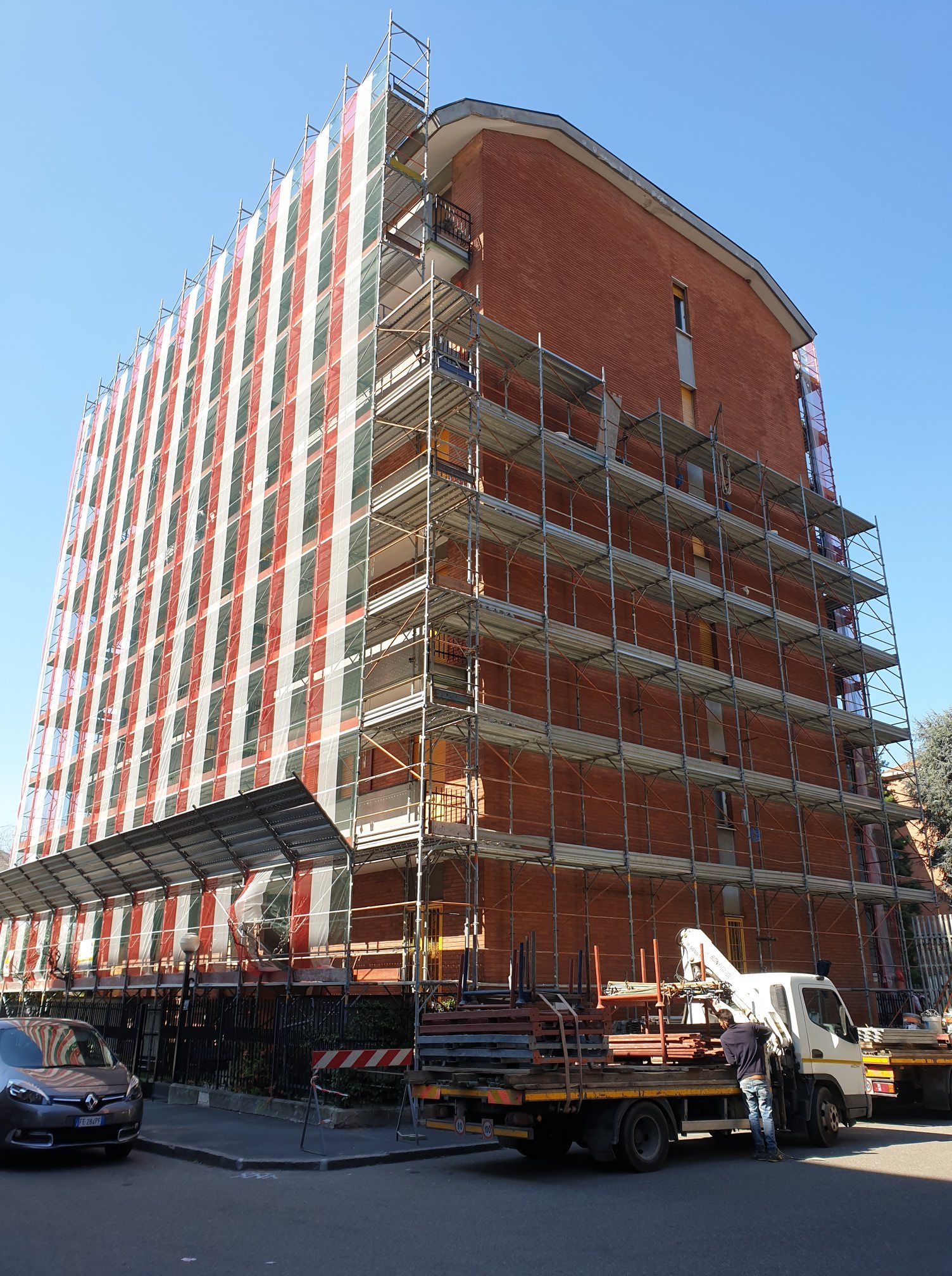 Edificio in costruzione, impalcature, facciata in mattoni, coperta da teli, camion parcheggiato nelle vicinanze.